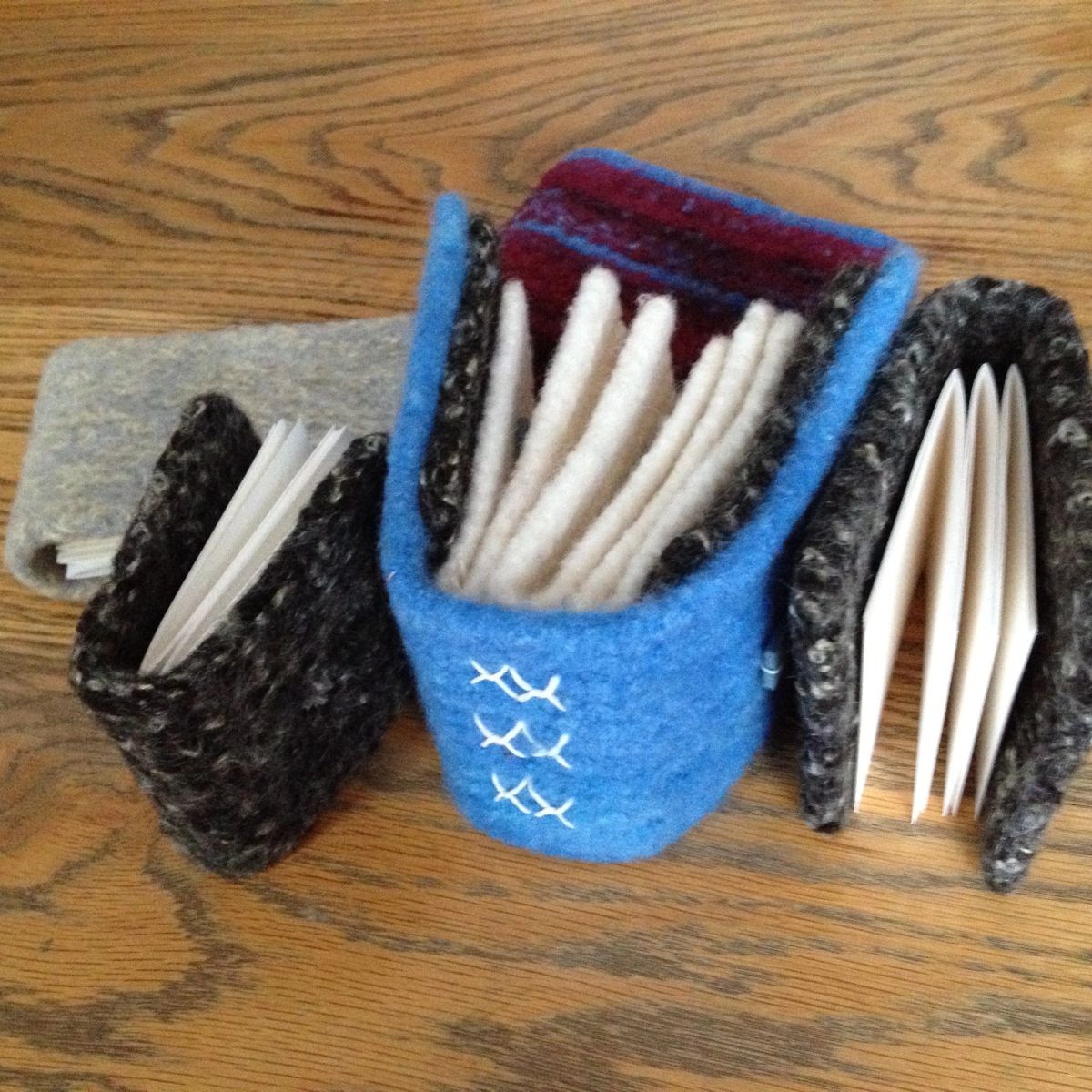 Three small books with nubbly boucle-textured covers in charcoal and blue rest on oak, alongside a blue felted vessel holding rolled cream-coloured spills. Three white cross-stitches mark the vessel's front.