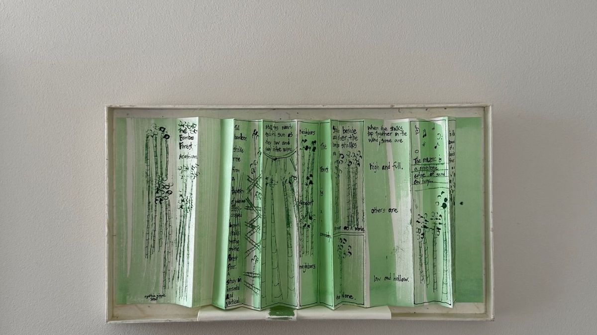 A white box holds a concertina of translucent green pages standing in zigzag folds. Fine ink drawings of bamboo stalks alternate with handwritten lines — "the music is a rustle", "high and full", "low and hollow".
