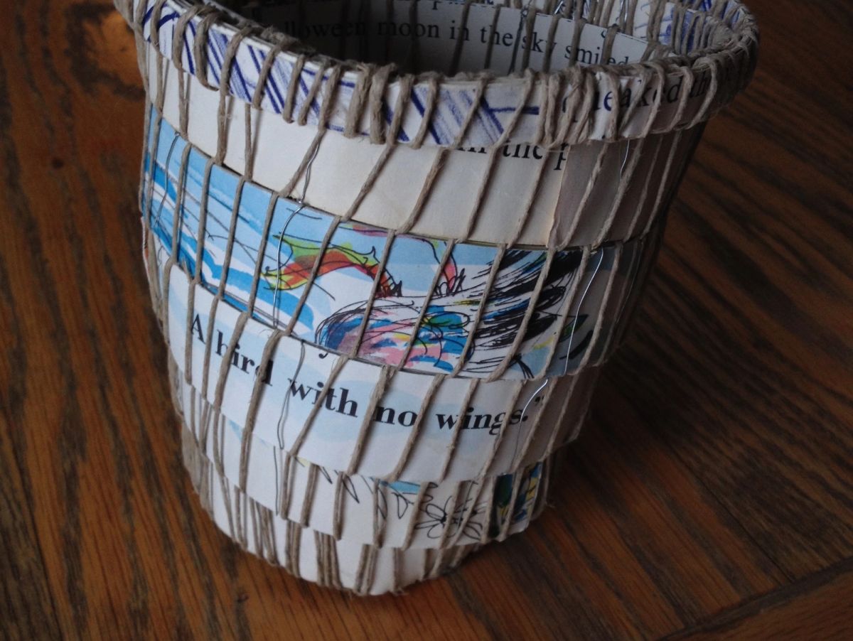 A tapered vessel woven from cut children's book pages over a warp of pale twine. Illustration fragments and text show between the bindings — "A bird with no wings", "Halloween moon in the sky smiled".