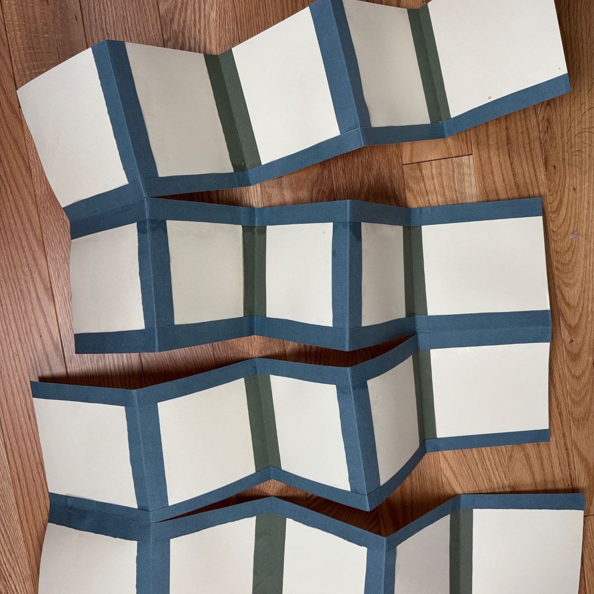 Several unbound concertina structures laid on oak, folded into zigzags. Each is faced with slate-blue and olive paper bands framing blank cream panels — the book form before anything is written in it.