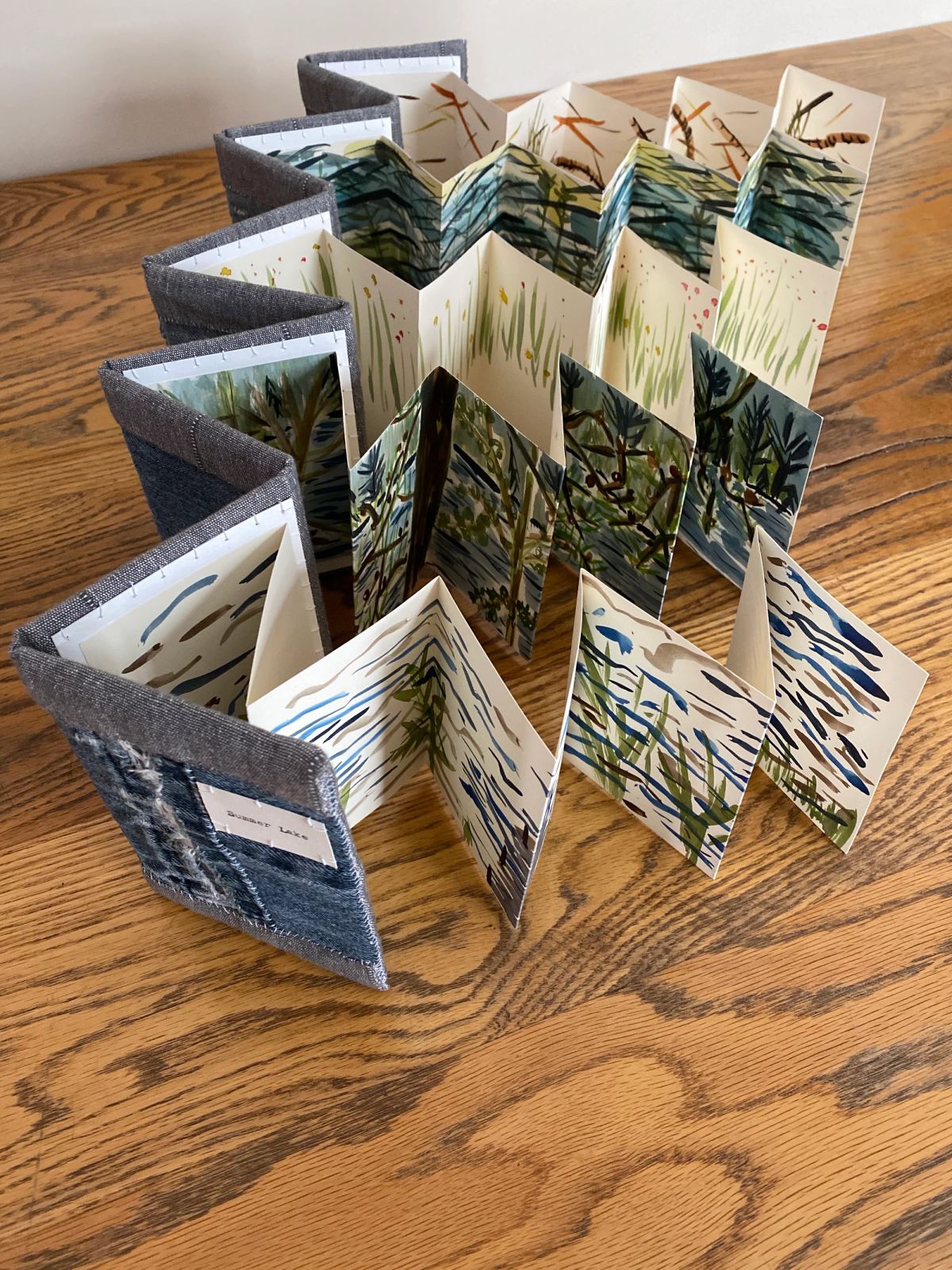 The same concertina book opened into a full circle, revealing painted panels of marsh grasses, reeds and dark pines above pale water. Loose watercolour strokes in green, blue and ochre run the length of the fold.