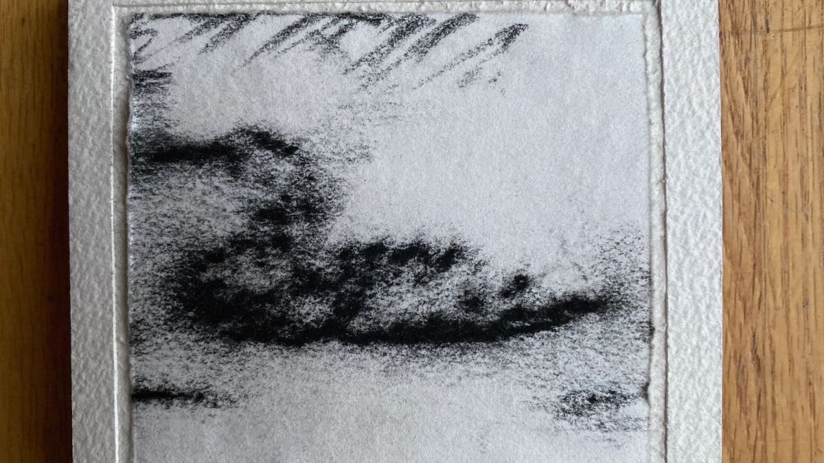 A small charcoal drawing on deckle-edged handmade paper, mounted on wood. A dense dark mass smudges across the lower half like a treeline seen through snow, with softly scraped diagonal strokes above.