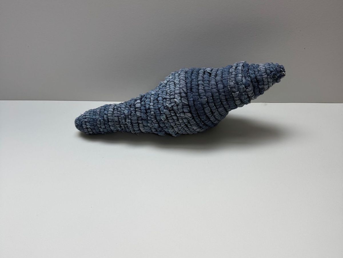 An elongated horn-shaped vessel coiled from denim-blue strips, tapering to a fine upturned point. The stitched rounds ripple along its length; it rests on white with a faint shadow.