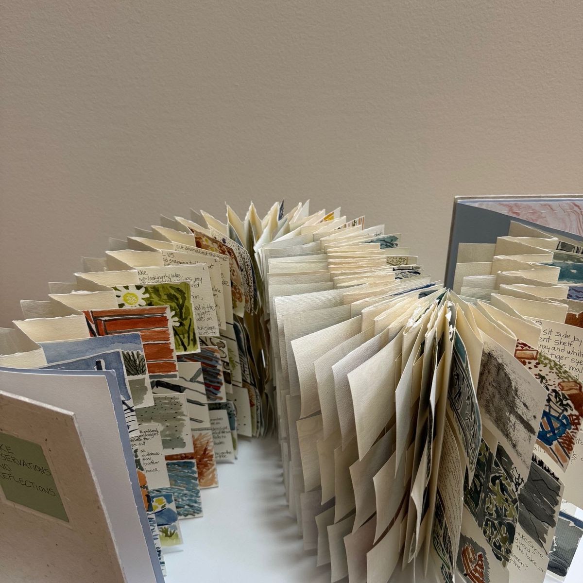 A large fan-bound book opens into a near-circle, hundreds of deckle-edged pages splaying outward. Each carries handwriting alongside small painted swatches — daisies, water, brickwork, weather notes.