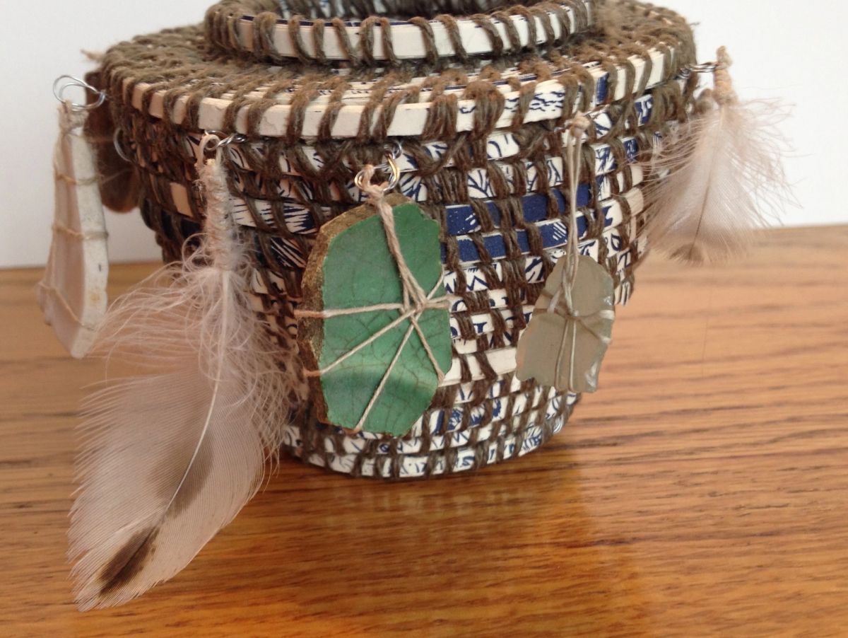 A lidded basket coiled from blue-and-white printed paper bound with coarse brown twine. Sea-glass shards in green and grey hang from wire loops alongside soft downy feathers.