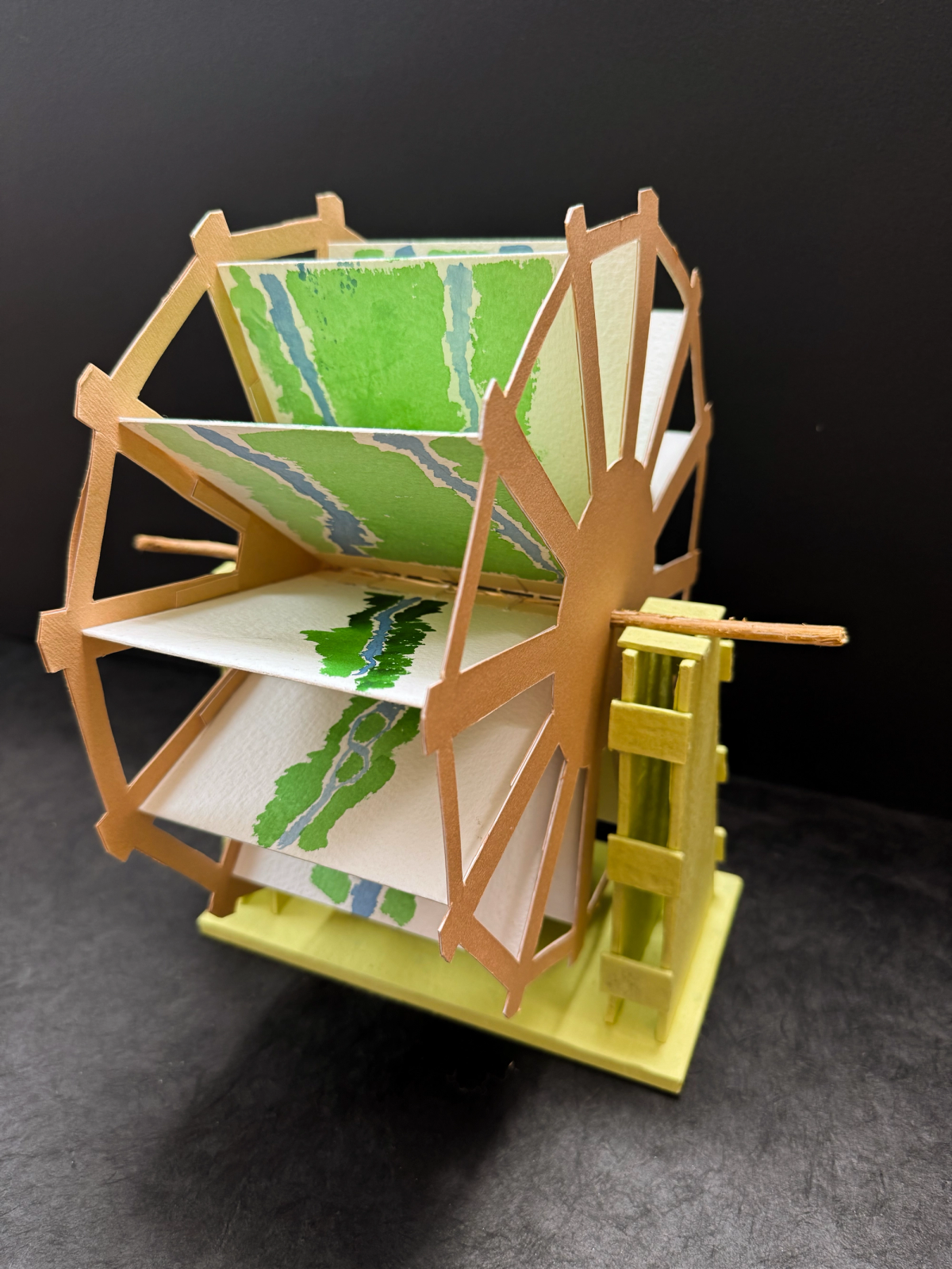 A three-dimensional kinetic book object shaped like a water wheel, crafted from light brown cardstock and set on a pale yellow base. Its rotating paddles hold textured watercolor paper panels displaying abstract green and blue river landscapes.