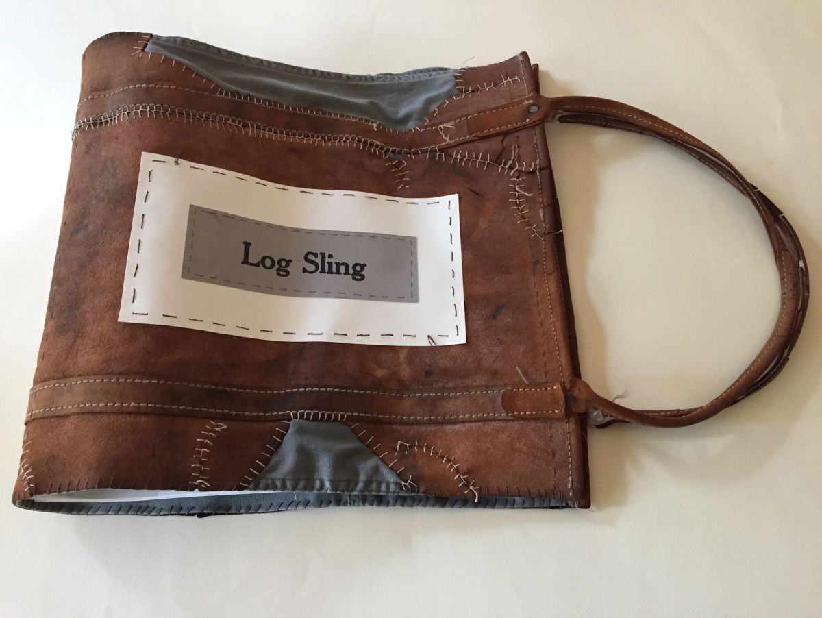 A book bound in worn brown leather with a carrying strap, its panels joined by coarse pale blanket stitching over grey fabric. A hand-stitched paper label reads "Log Sling".