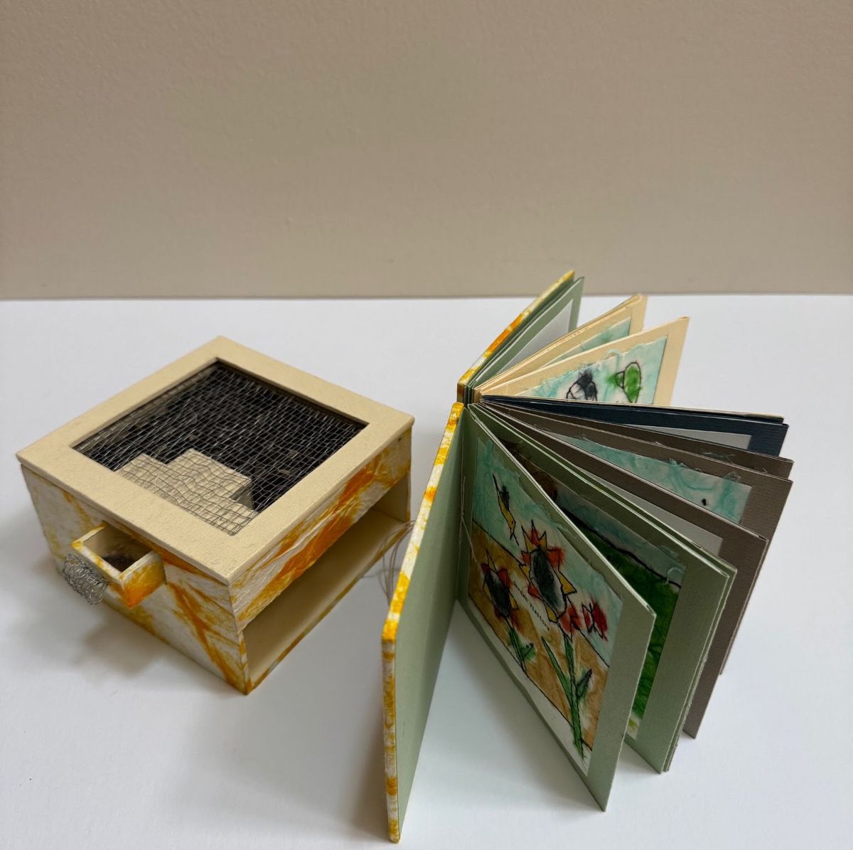 A cream-painted wooden box with a dark mesh window and a small drawer sits beside a book fanned open. The pages hold childlike drawings of sunflowers and bees in watercolour and crayon.