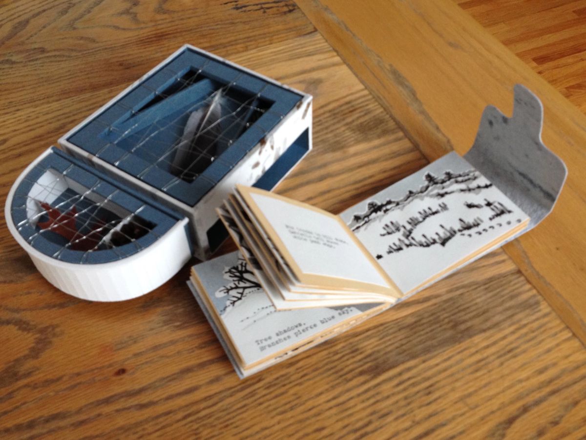 A blue and white clamshell box with a latticed lid sits open on oak boards beside a small book unfolding into a cut-profile page. Inside, ink drawings of bare winter trees run along a snowbank horizon.