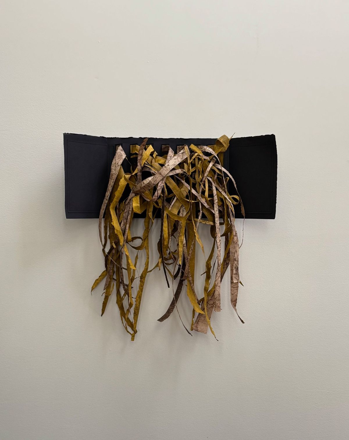 A wall-mounted piece: a matte black panel with a dense curtain of torn paper strips falling from its lower edge in ochre, gold and mottled bark-brown. The strips curl and twist as they hang, several dropping well below the panel.