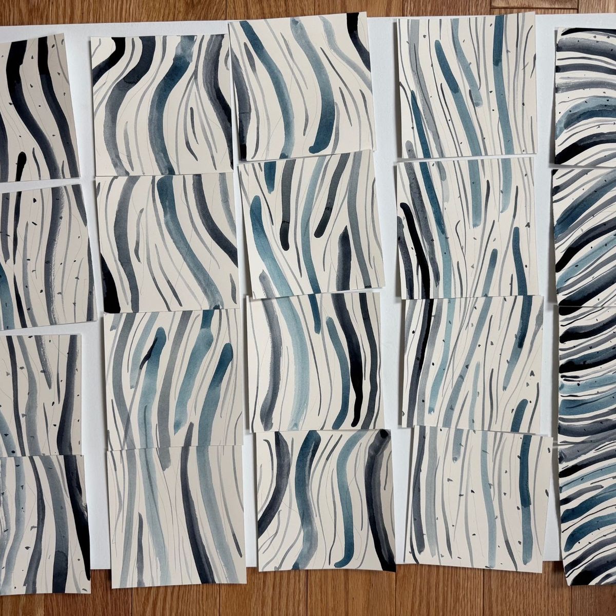 A grid of small watercolour studies laid on oak. Each shows vertical wavering lines in indigo, slate and pale blue on cream paper, like reeds or reflections; the rightmost sheet curves into concentric arcs.