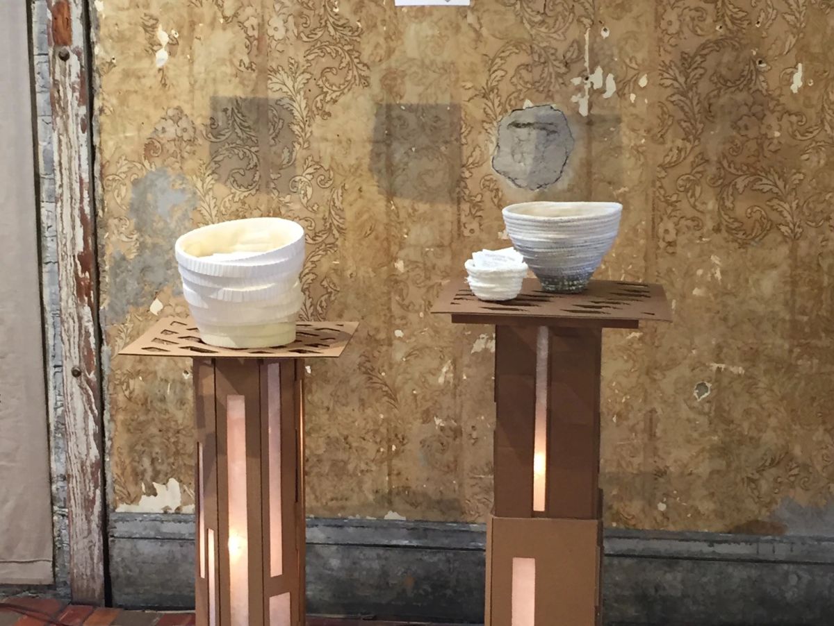 Two pale coiled paper vessels on slim wooden plinths lit from within, in a gallery with peeling floral wallpaper and bare plaster. Warm light spills through cut slots in the pedestals.