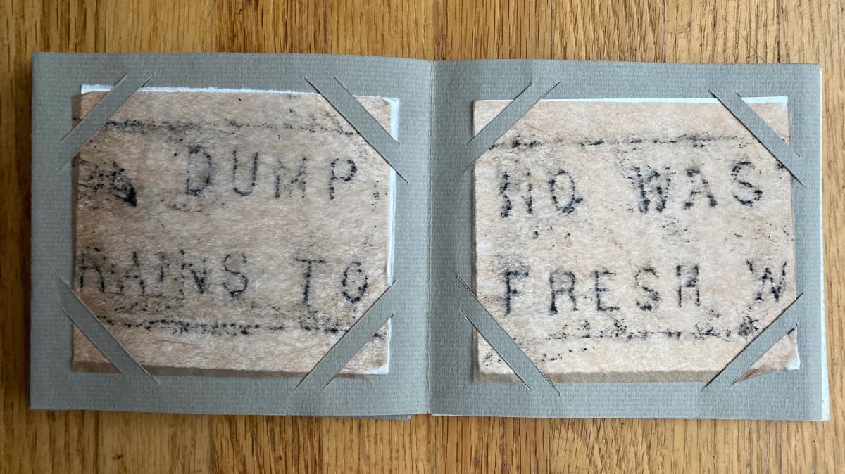 A grey book lies open on oak, each page holding a slab of coarse pulped paper bearing transferred fragments of stencilled type — "DUMP", "RAINS TO", "NO WAS", "FRESH W" — held by grey corner tabs.