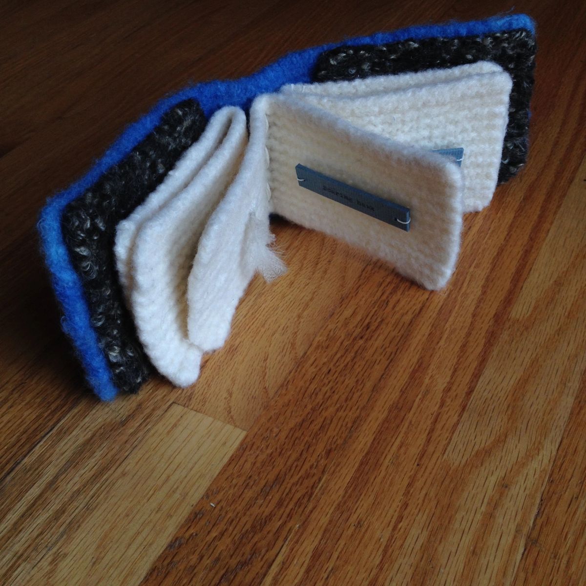 Felted books lie open on an oak floor, cobalt and charcoal covers framing thick knitted cream pages. A small grey printed label is stitched to one page. Raking light picks up the loose fibres.