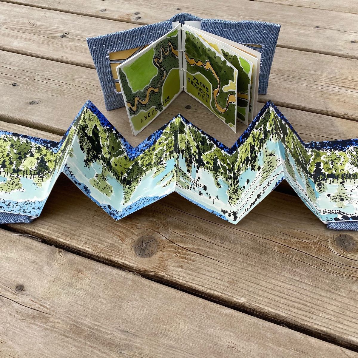 Two accordion books on weathered decking. The foreground book zigzags into a painted panorama of dense green forest above pale blue water; behind it a smaller book shows a winding river map lettered "ANCESTORS" and "FLOWS THROUGH OUR".