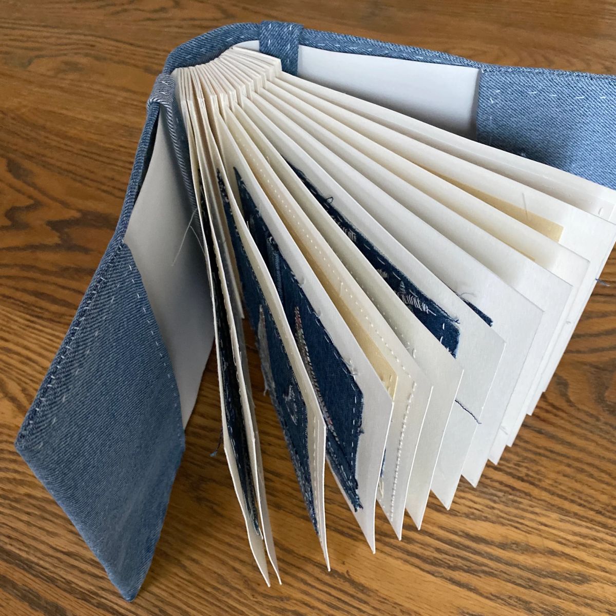 A denim-covered book fanned open on oak, its cream pages interleaved with ragged indigo denim shapes. White running stitch crosses the pages; loose threads catch the light.