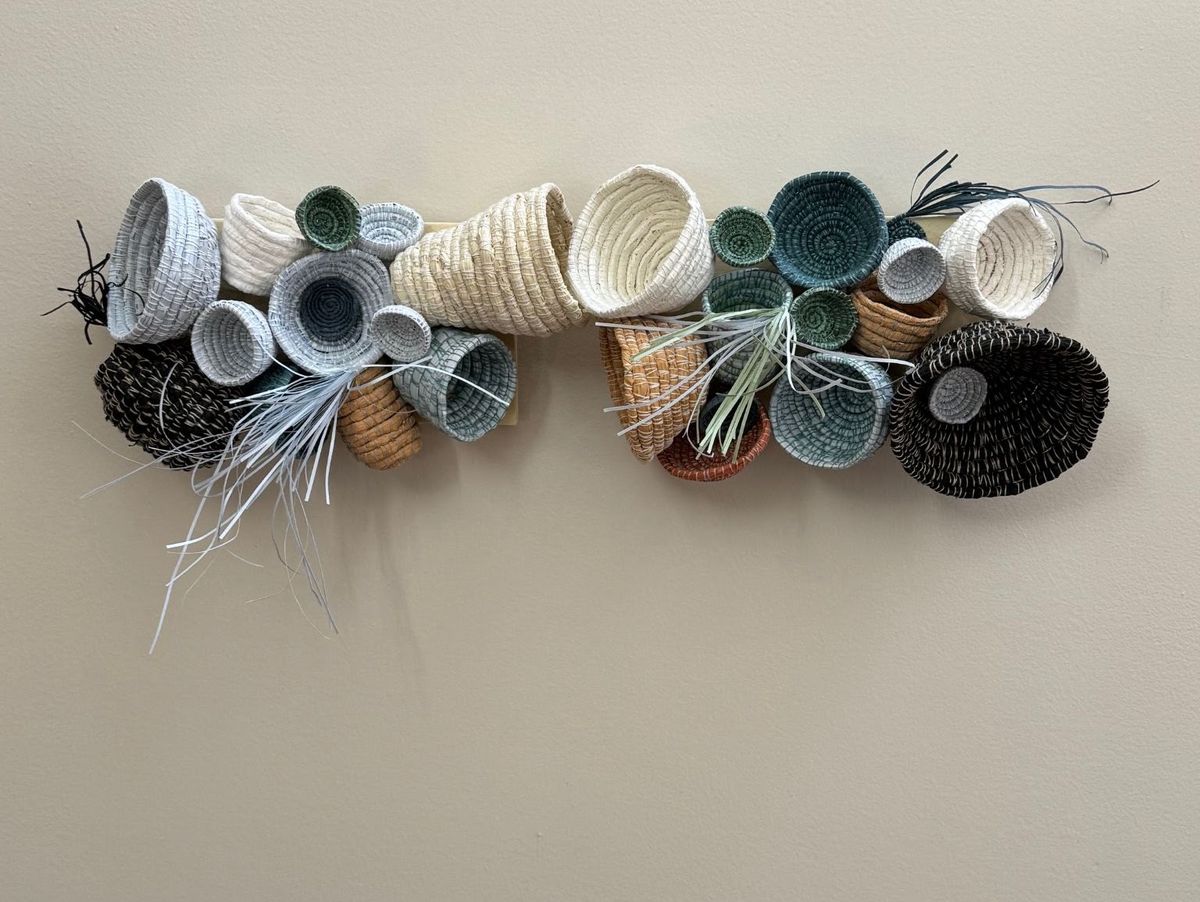 A wall-mounted cluster of coiled baskets on a pale batten, ranging from open bowls to closed cones in white, sage, rust and black. Sprays of thin pale and dark grasses burst from between them.