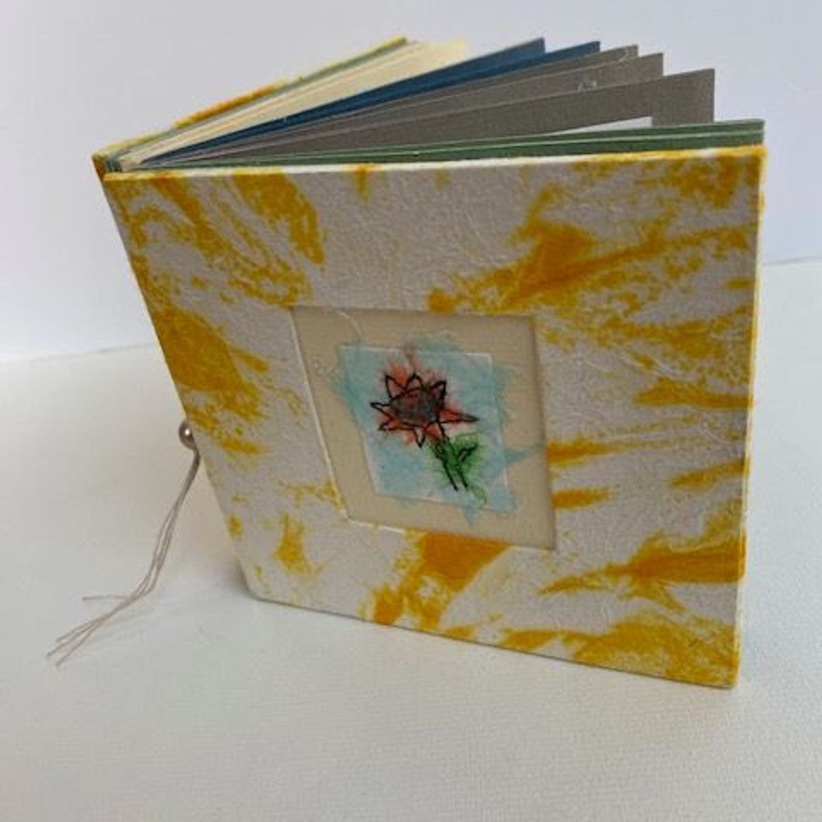 A small book stands on white, its cover papered in white and marigold-yellow marbling. A recessed window frames a stitched sunflower on pale aqua. Coloured page edges show at the top; a thread tie hangs loose.