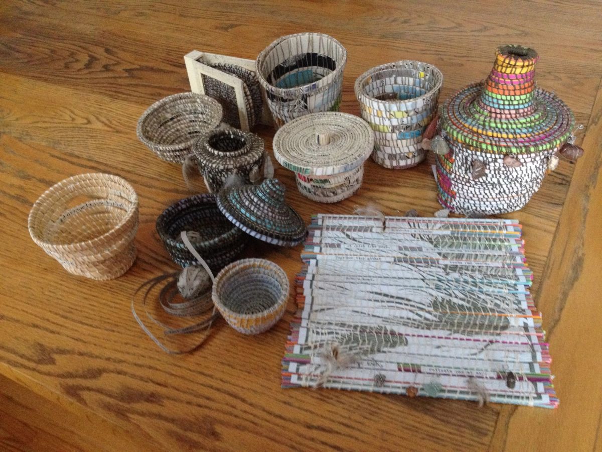A gathering of coiled paper vessels on an oak floor — open bowls, lidded jars, a tall rainbow-banded pot — most worked in newsprint greys with fragments of type showing. A woven mat with feathers and shells lies in front.