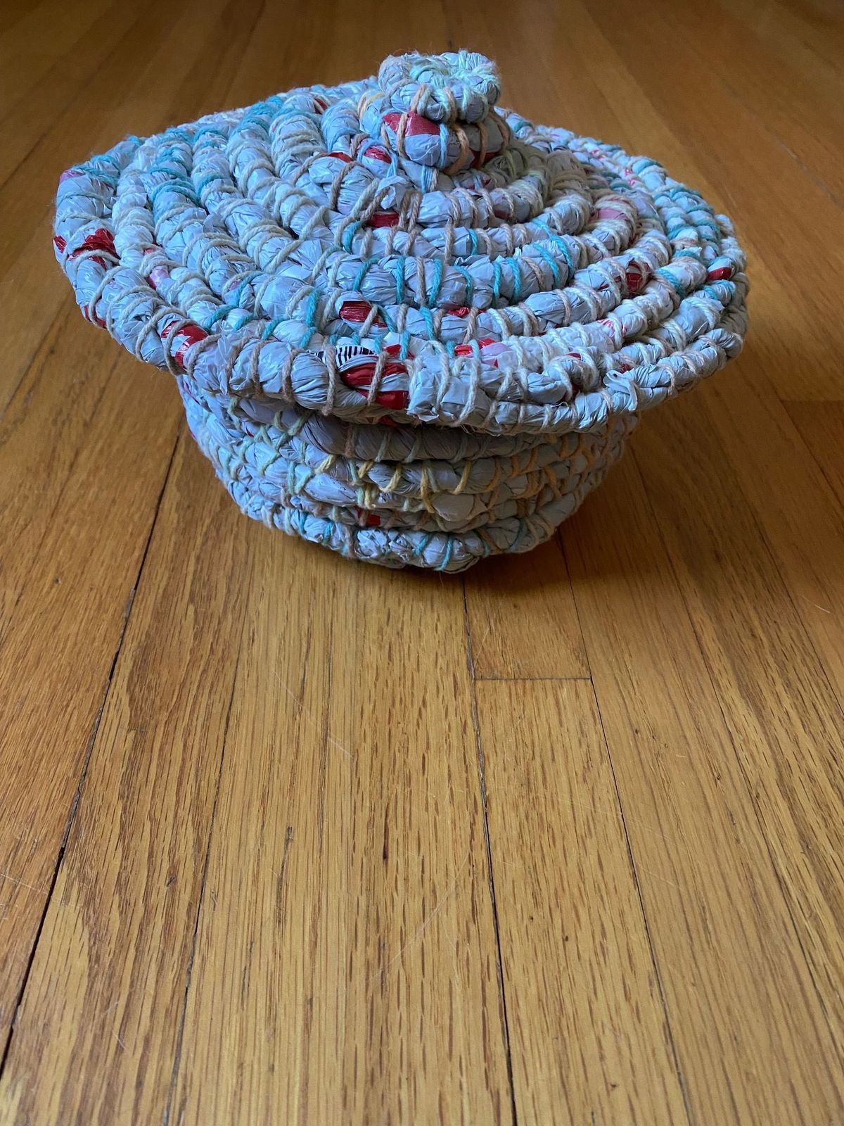 A lidded coiled basket on oak, its wrapped strips pale cream flecked with turquoise and scarlet. The domed lid spirals to a knob; the binding thread shows as fine diagonal stitching throughout.