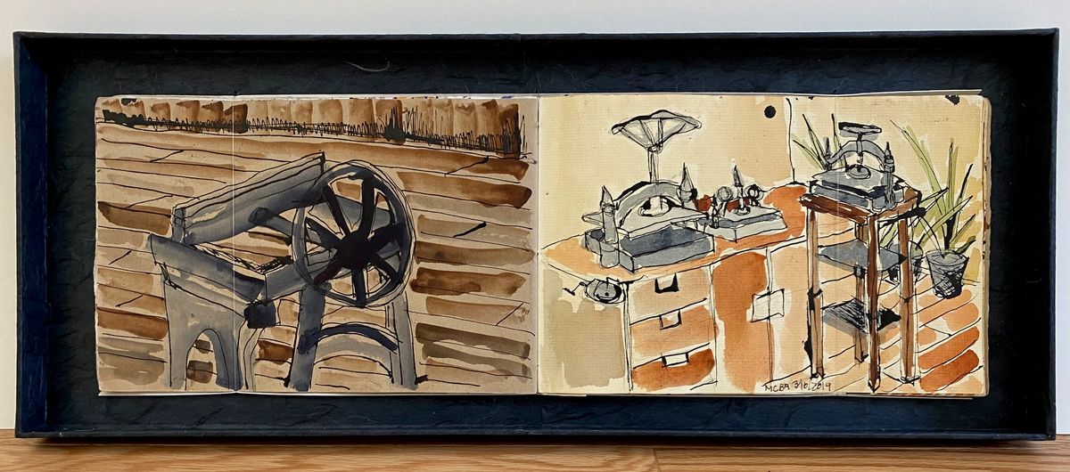 A sketchbook lies open inside a deep black shadow box. Two ink and wash drawings in sepia and rust: a spoked wheel and timber frame on the left, a cluttered workbench with tools and a potted plant on the right.