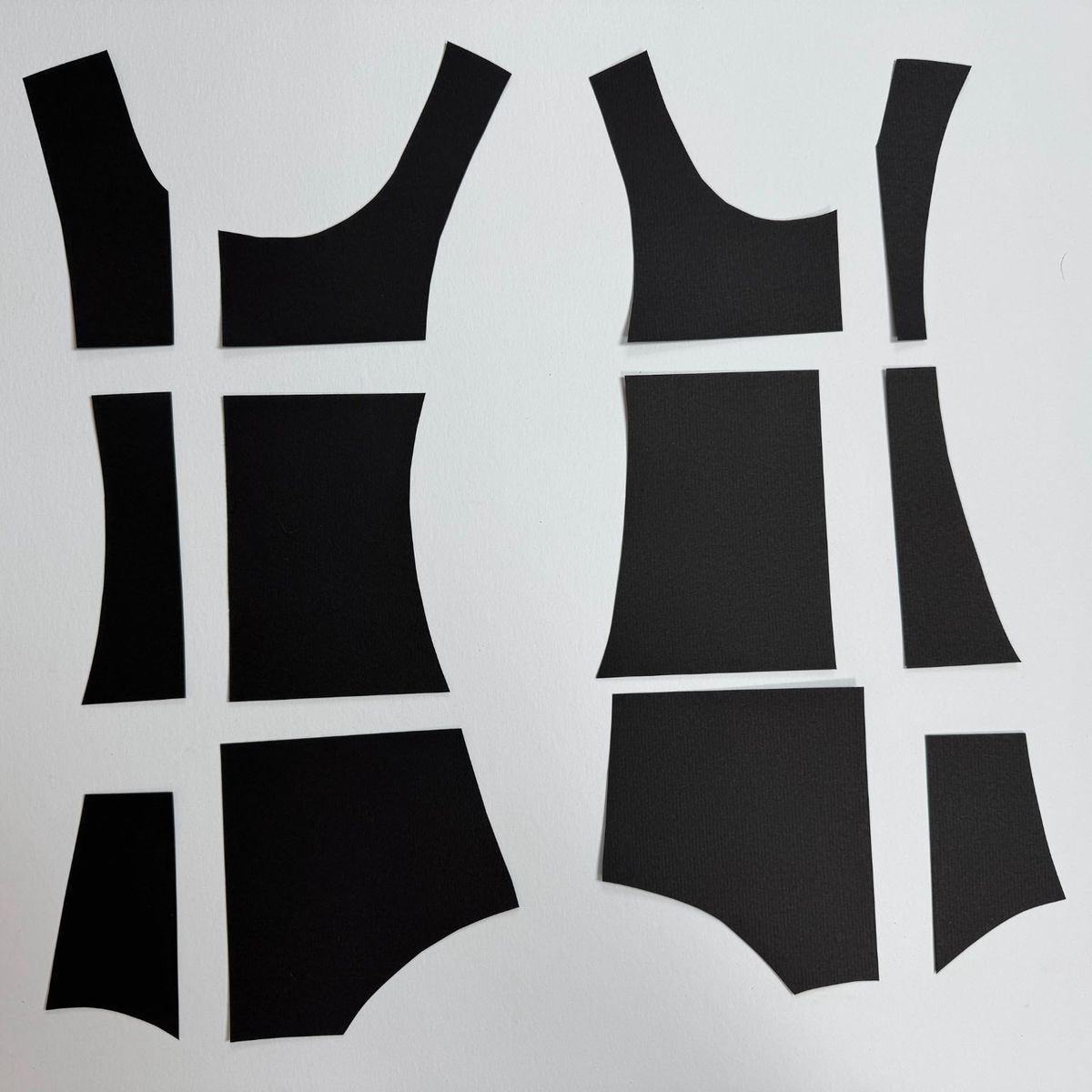 Twelve pieces of matte black paper arranged in two columns on white, cut into curved garment-pattern shapes — bodice panels, straps, gently flared skirts. Crisp edges throw thin shadows.