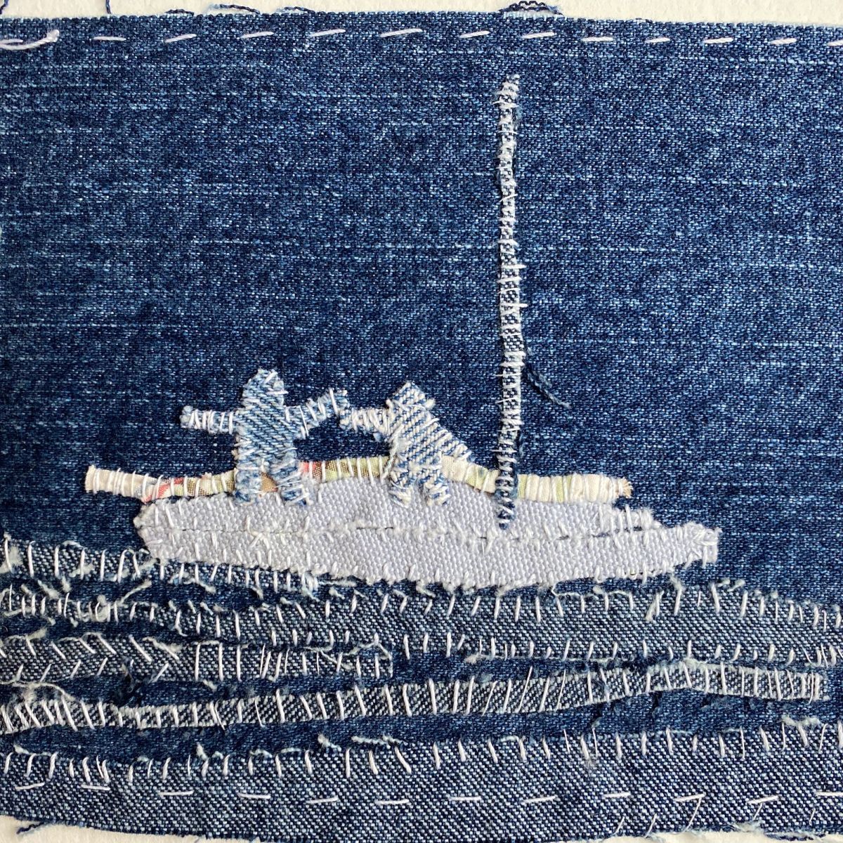 A sailboat built from layered scraps of indigo denim, hand-stitched onto a denim ground with white running stitch. The hull is pale twill, the mast a dense column of white thread, the water a stack of frayed strips.