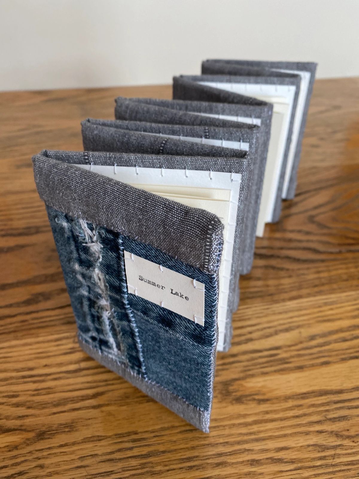 A concertina book stands zigzag on oak, its covers pieced from faded denim and coarse grey linen. Cream and white pages show between the folds. A typed label reads "Summer Lake".