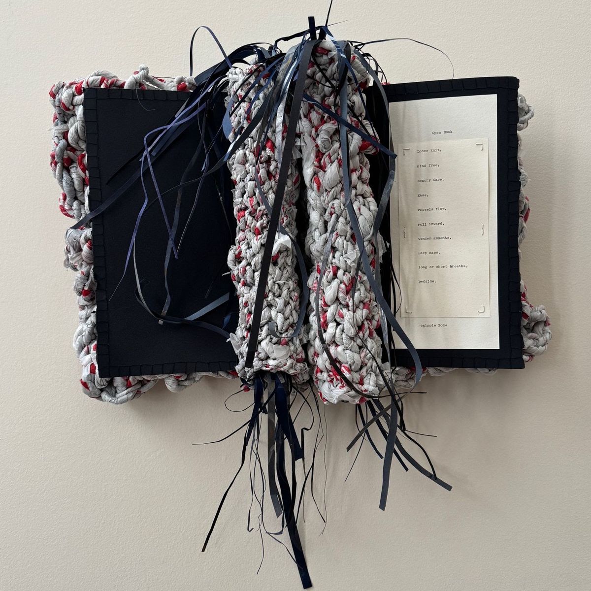A book-shaped wall piece crocheted from pale rag strips flecked with red, mounted over black board. Navy ribbons spill from the centre like a spine. The right page holds a typed poem beginning "Loose knit. Mind free."