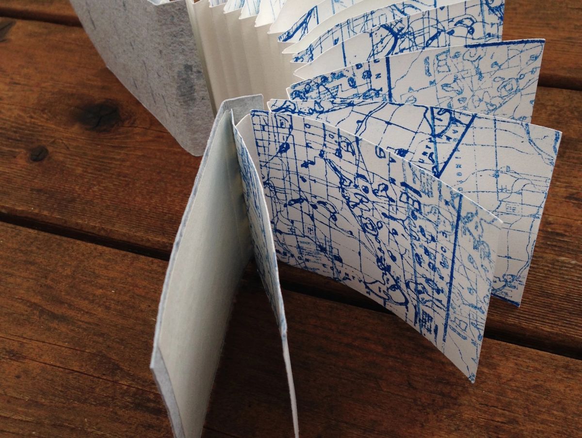 A small concertina book on weathered wood, its deckled pages printed edge to edge with blue survey-map linework — contours, section grids, lake outlines. Grey handmade-paper covers stand behind.