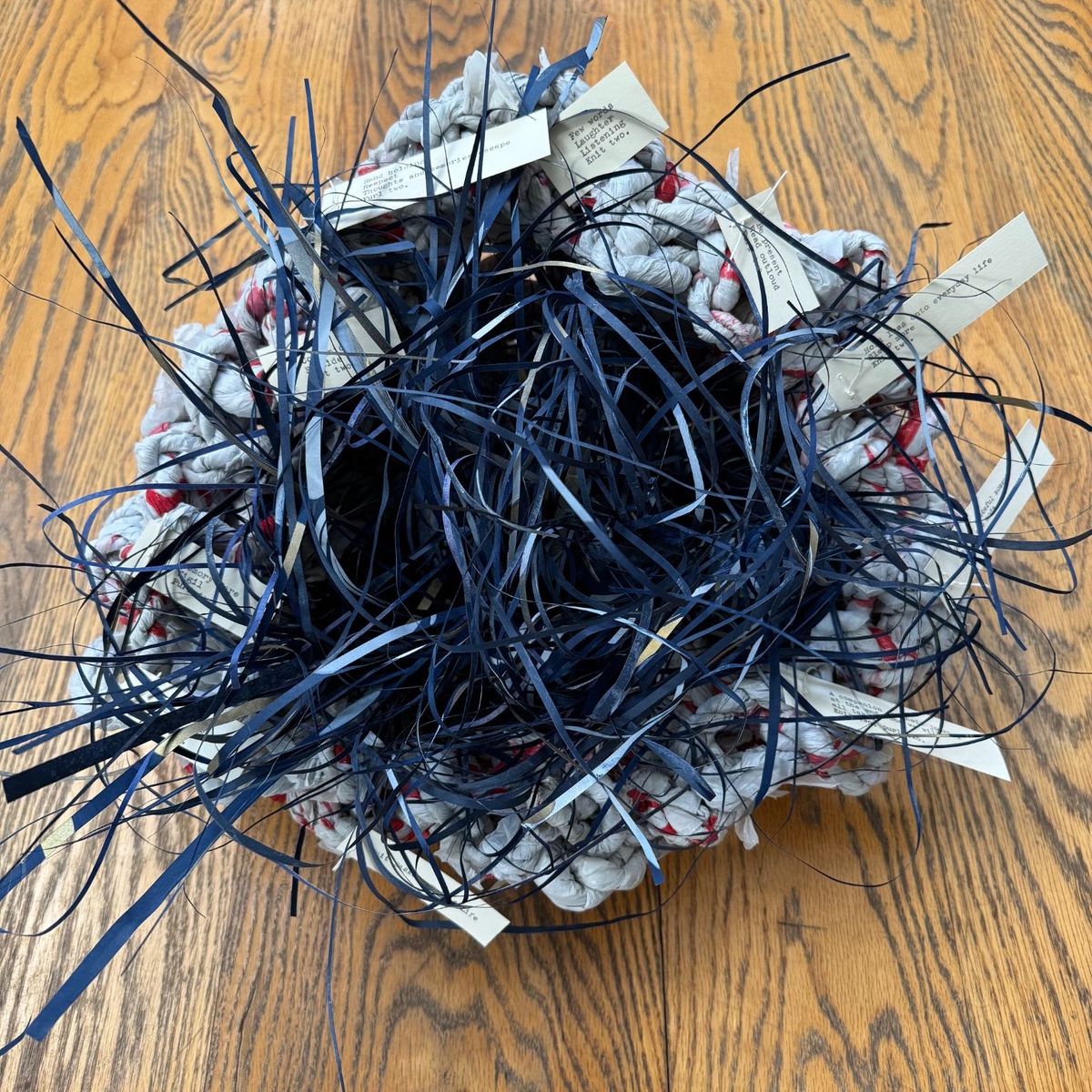 Overhead view of a coiled basket crocheted from pale rag strips flecked with red, its centre erupting with a tangle of navy raffia. Small typed paper tags are tucked between the coils.