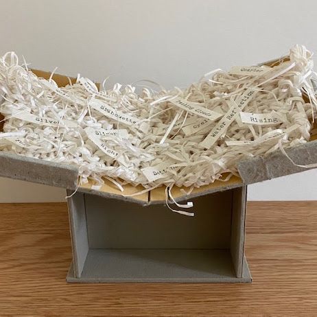 A grey board cradle supports a shallow woven bowl of cream paper strips. Typed word-slips are worked into the weave — "Silver", "Rising", "Water Reflection", "Passing Clouds", "Quarter" — with curled ends lifting free.