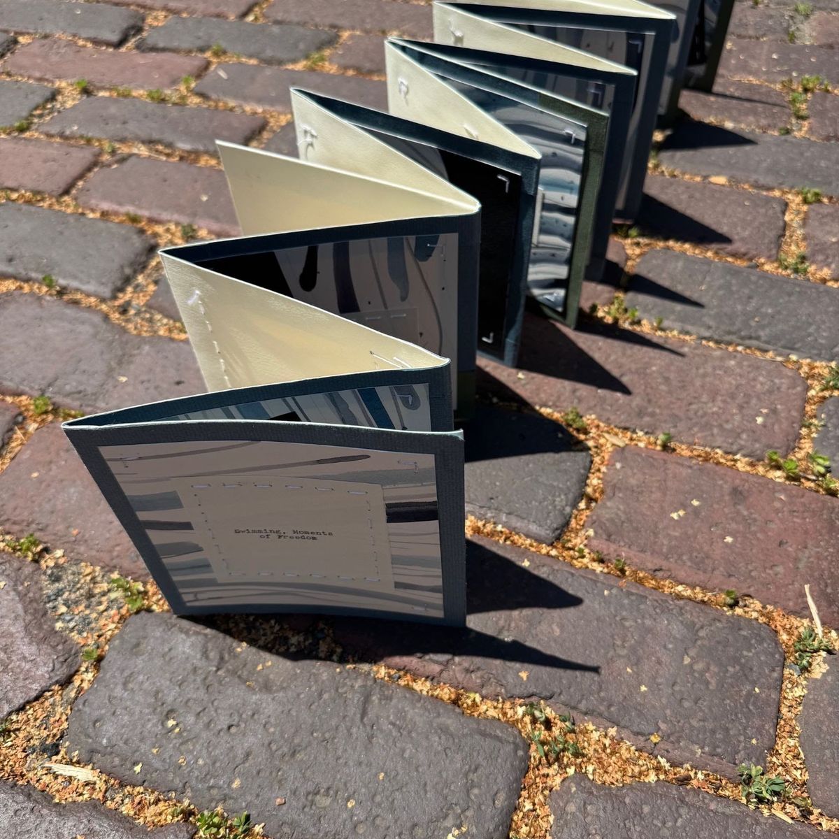 An accordion book stands on sunlit cobbles, casting long shadows. Dark blue covers open onto pale silver-grey pages printed with soft blurred forms. A typed label reads "Relaxing, Moments of Freedom".