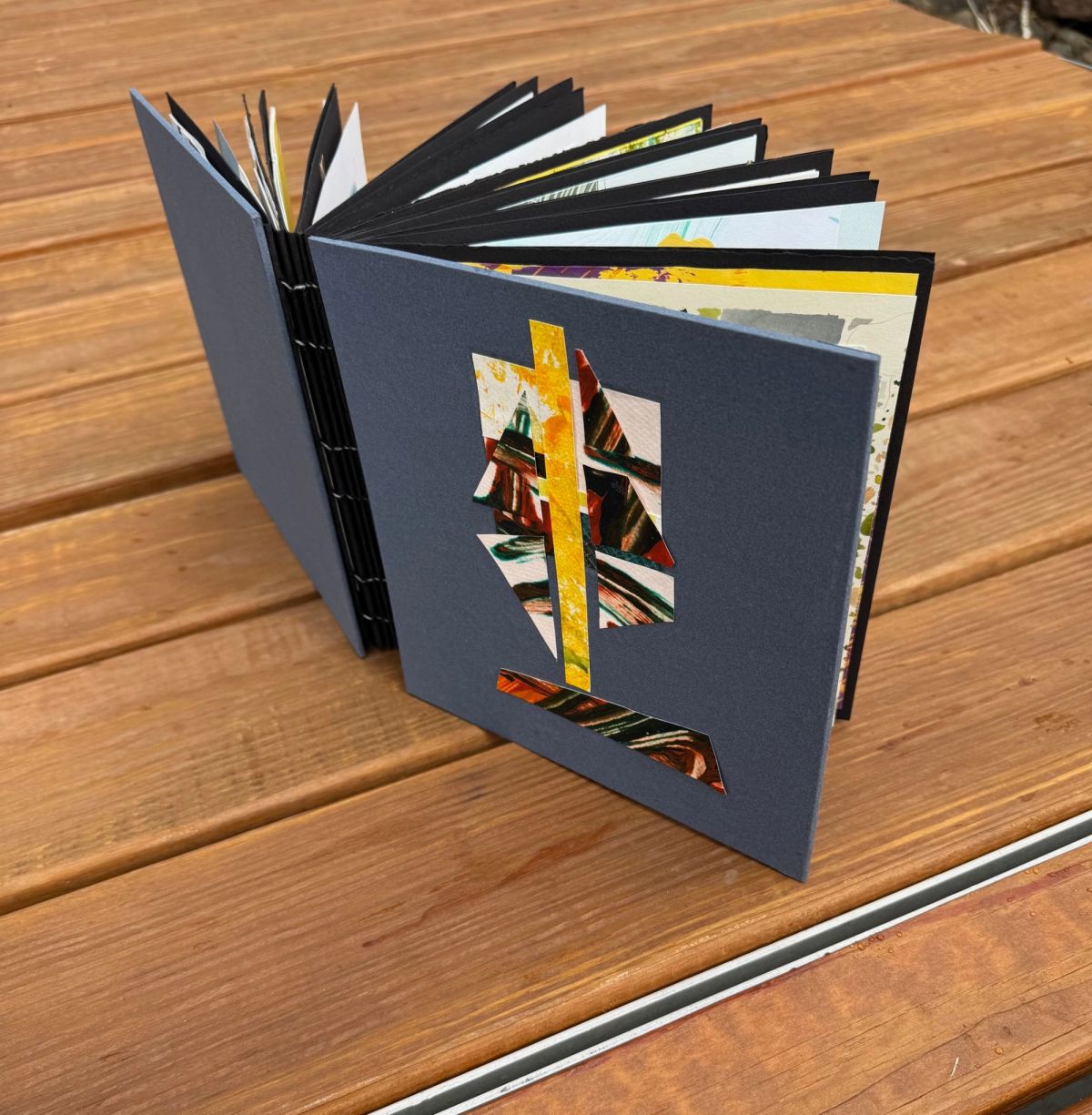 A slate-grey artists' book stands open on weathered decking, its exposed spine stitching visible. A window cut through the cover reveals collaged painted papers underneath — a yellow stripe crossing marbled green and rust.