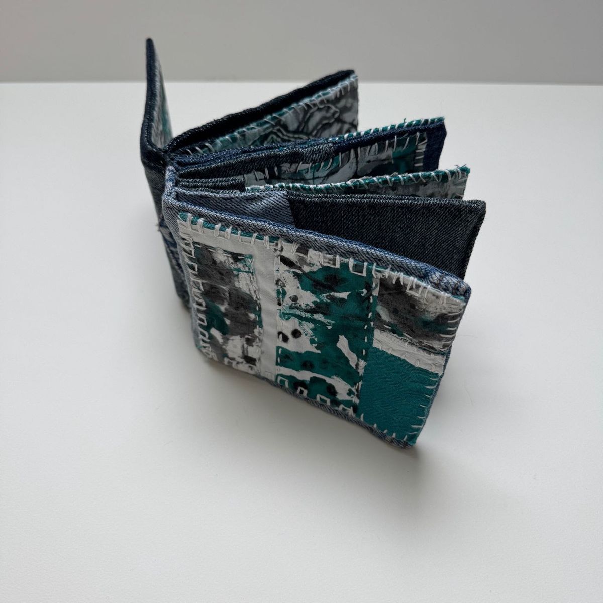 A small accordion book standing on white, covers pieced from indigo denim. Its pages carry blocky printed marks in teal, black and white, edged with dense blanket stitch in pale thread.