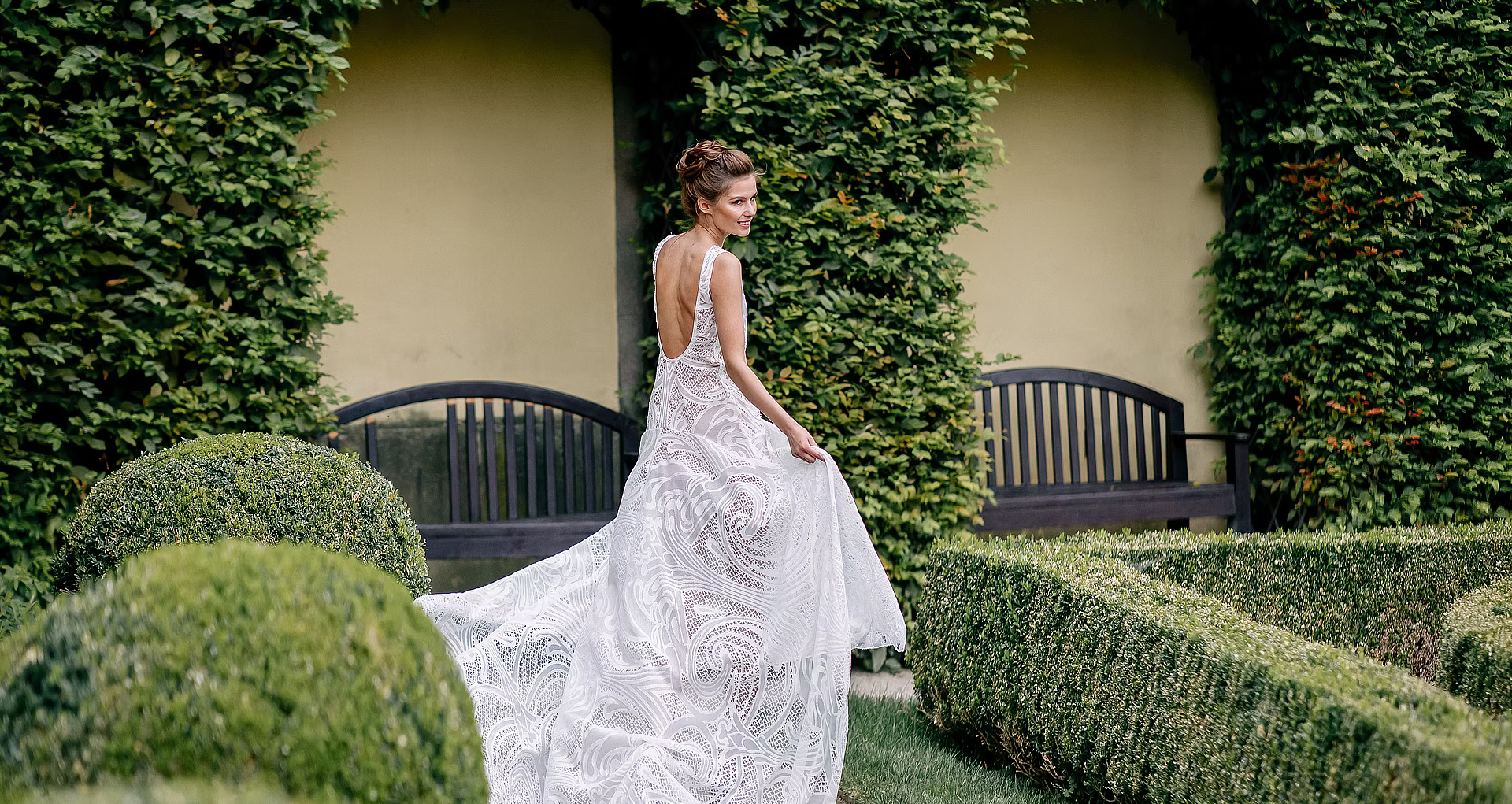 Bride in a white lace gown with an open back, looking over her shoulder in a sunlit garden with trimmed hedges and an ivy-covered wall