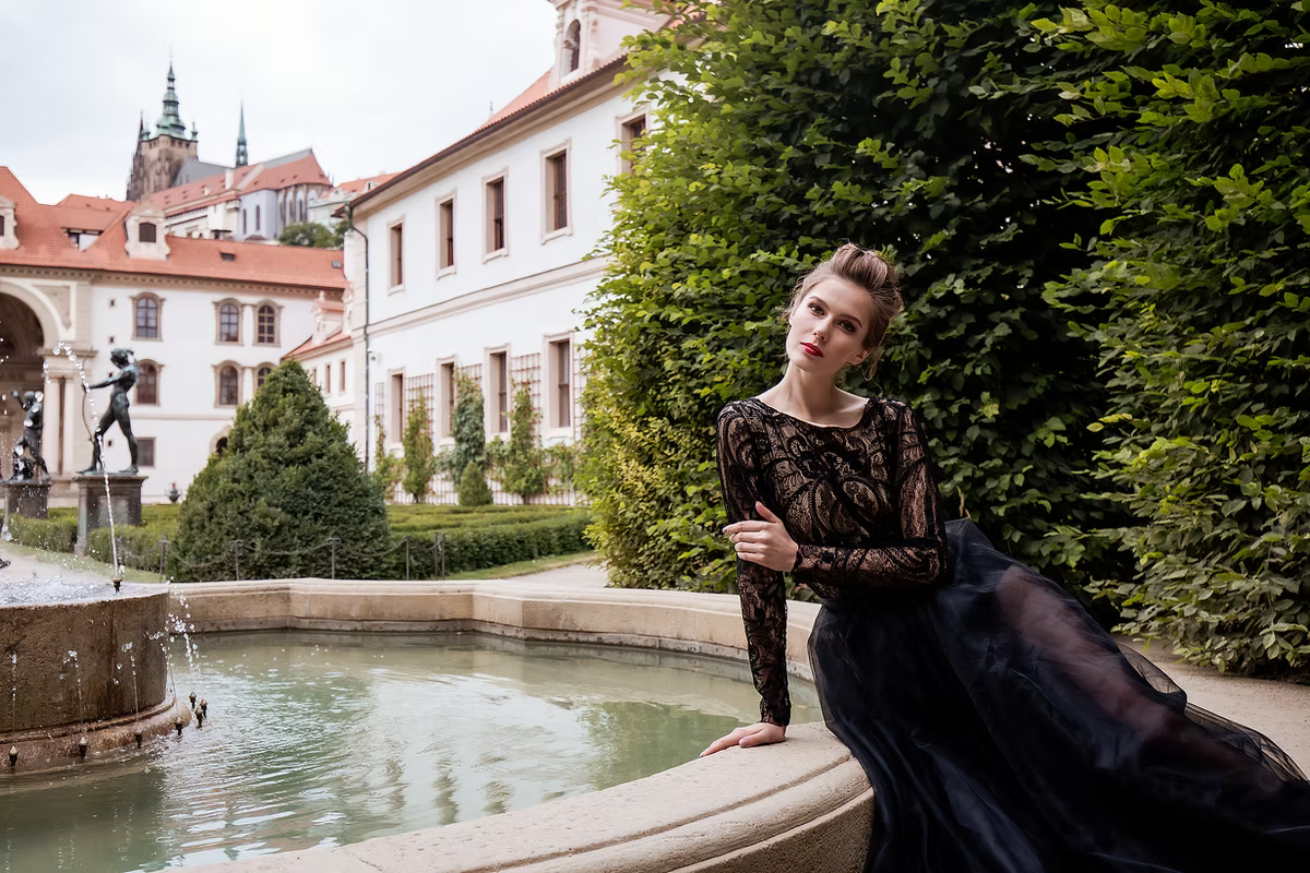Model in a black evening gown with a lace bodice and flowing tulle skirt, standing in a dramatic garden courtyard with cascading foliage