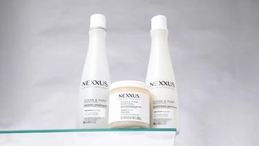 The Nexxus Clean & Pure collection, including Shampoo, Conditioner, and Scalp Scrub, is shown standing on a glass shelf against a white marble background.