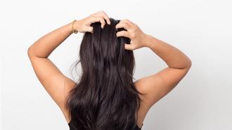 Woman with Dark hair facing a grey wall with both of her hands messaging her scalp.
