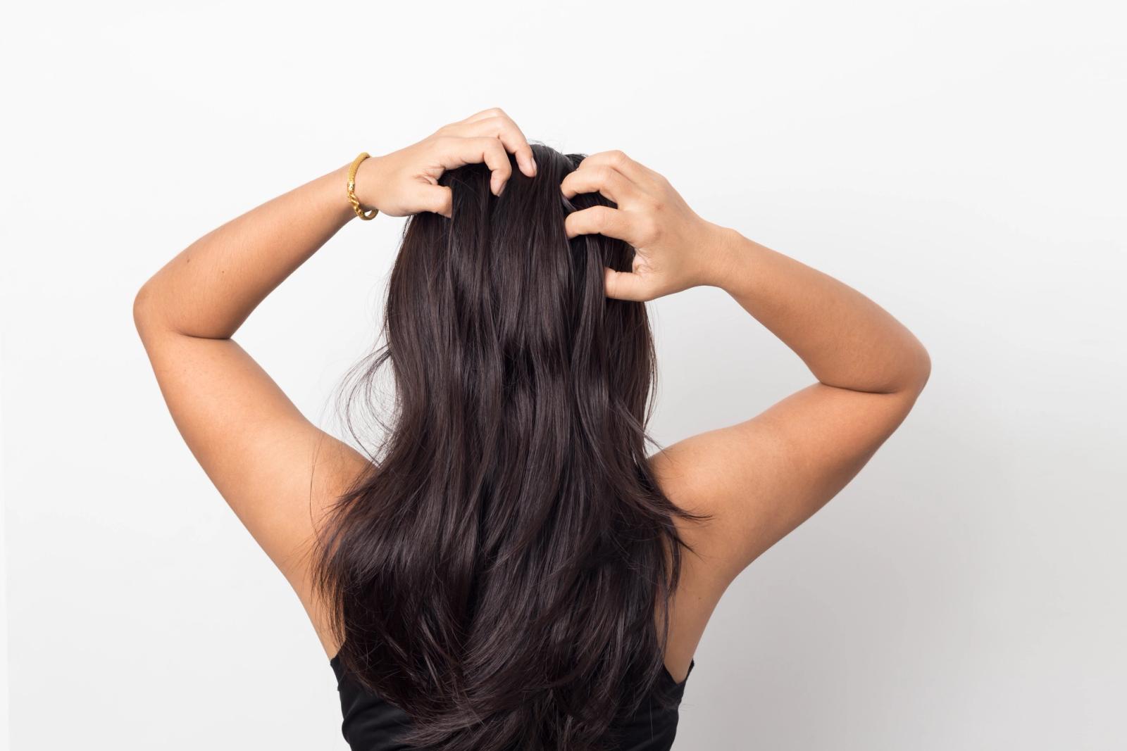 Woman with Dark hair facing a grey wall with both of her hands messaging her scalp.