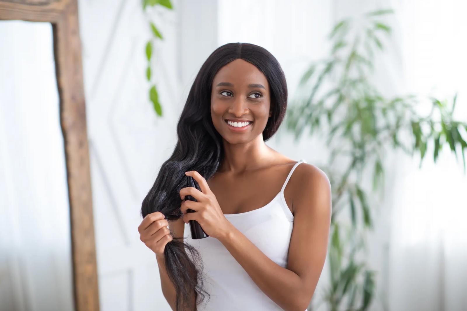 Woman wearing a white tank top smiles whilst spraying product into her long black hair