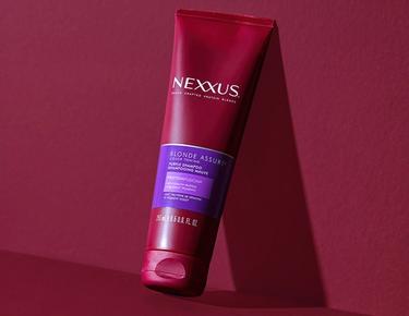 Nexxus Blonde Color Assure Shampoo leans again a dark red background.