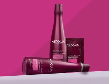 The Color Assure collection, including a bottle of shampoo, a bottle of conditioner, and a hair mask packet, is show against a pink background.
