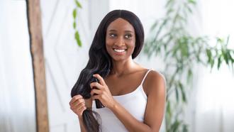Woman wearing a white tank top smiles whilst spraying product into her long black hair