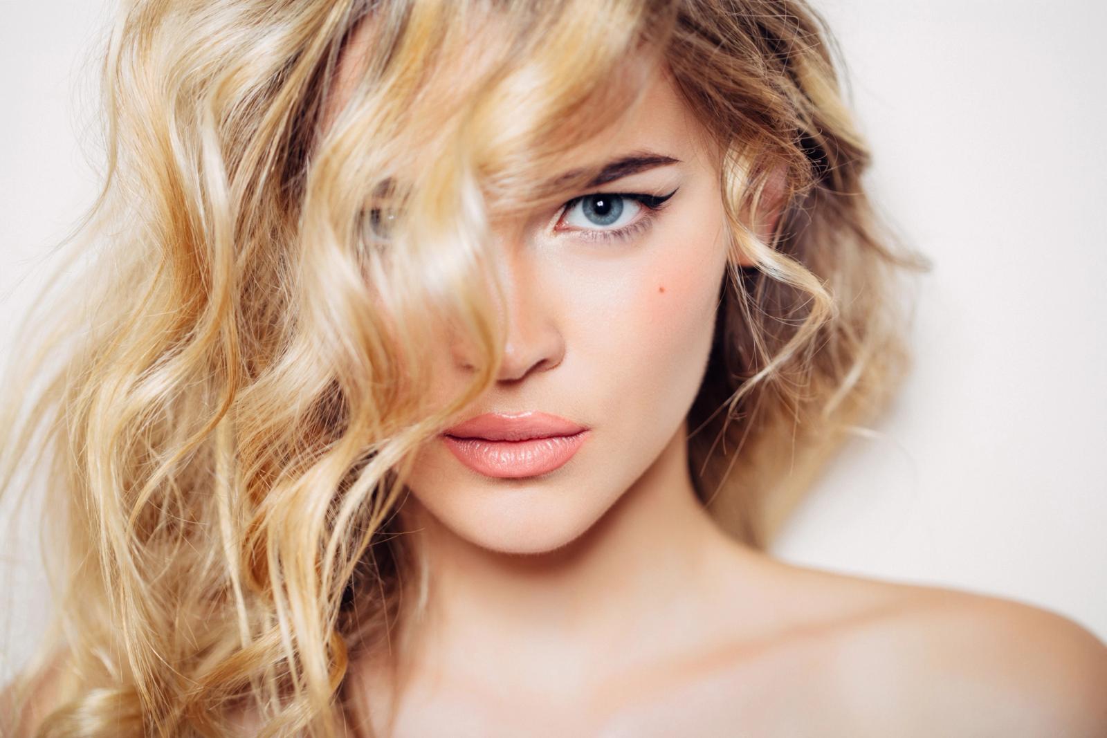 woman with wavy blonde hair leaning against the white wall with a couple of strands of hair are covering her face.