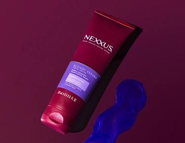 A bottle of Nexxus Blonde Color Assure Shampoo is shown lying next to a dollop of purple shampoo against a maroon background.