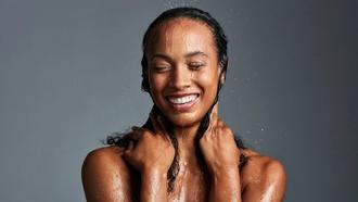 Woman faces the camera smiling. water is falling on her head as she holds her hair at her neck with both hands.