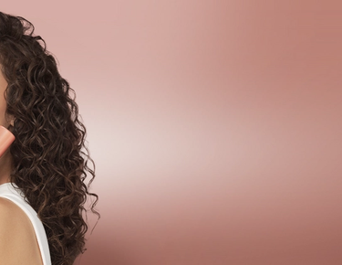 A model with dark curly hair is shown against a rose-gold background while holding a can of Nexxus Volume Medium Hold Mousse.