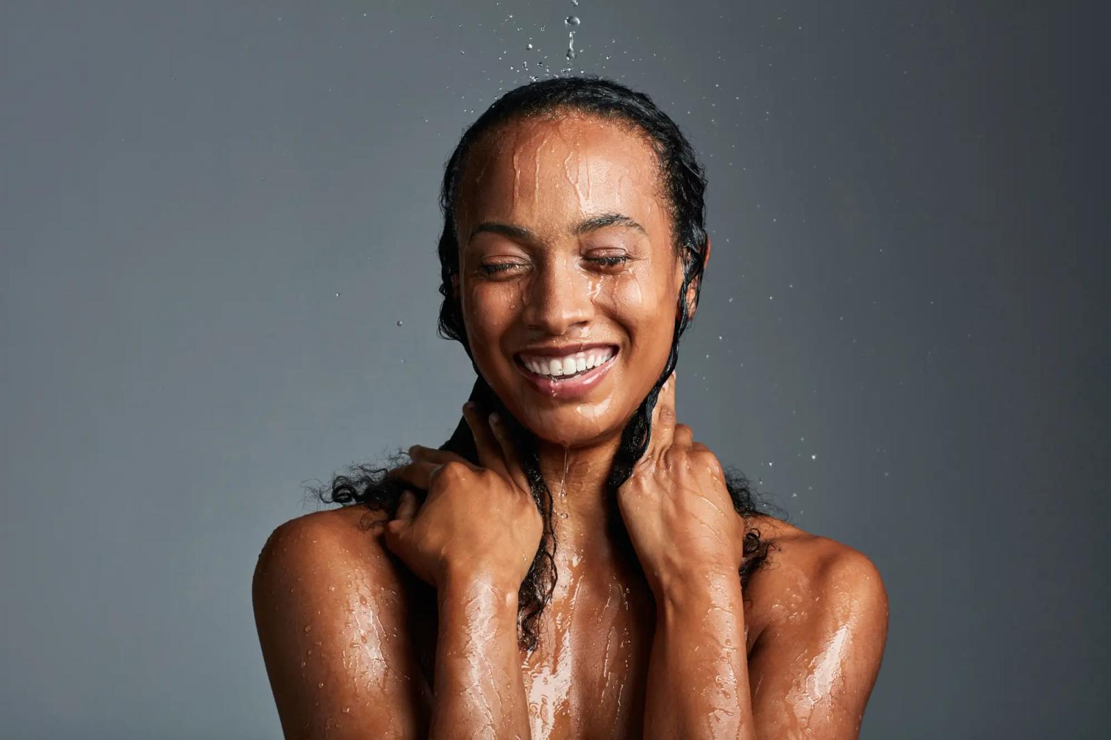 Woman faces the camera smiling. water is falling on her head as she holds her hair at her neck with both hands.