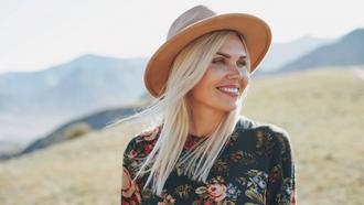 Woman with blonde hair in a beige hot standing in a field in the mountains smiling and looking out to the views.